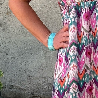 A woman stands outdoors against a textured concrete wall wearing a short-sleeved midi dress with a vibrant pink, purple, and blue floral pattern. She is wearing black ankle boots and standing next to a black planter with green leaves.
