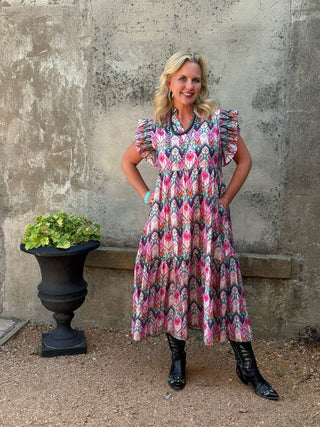 A woman stands outdoors against a textured concrete wall wearing a pink, black, and white patterned midi dress with ruffled sleeves and black boots.