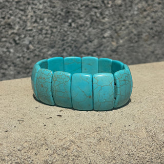 A turquoise stone segmented bangle bracelet with natural matrix veining, resting on a textured concrete surface against a gray stone wall background.