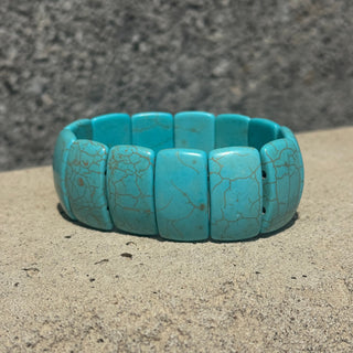 A turquoise stone beaded bracelet with natural crackle matrix, resting on a textured sandy surface with a blurred gray stone background.