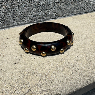 A dark tortoiseshell bangle bracelet with gold-tone metal studs, resting on a light concrete surface against a textured gray wall.