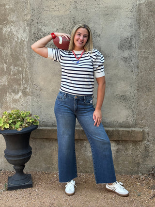 A woman stands against a textured concrete wall wearing a black and white striped short-sleeve t-shirt, blue wide-leg jeans, and white sneakers, holding a football to her head.