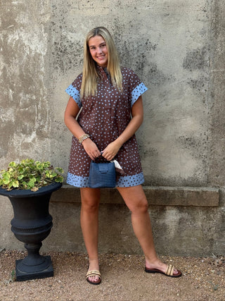 A woman stands outdoors against a textured concrete wall wearing a brown short-sleeve mini dress with a white floral pattern and light blue polka-dot trim. She is holding a small blue handbag and wearing flat sandals.
