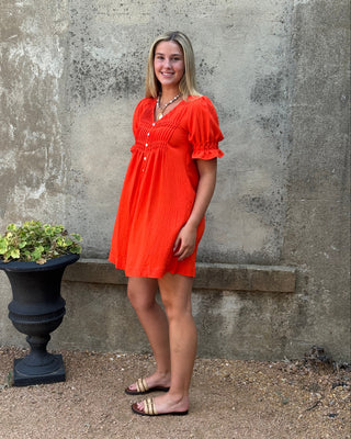 A woman modeling a bright orange short puff-sleeve dress with a smocked bodice and button-front neckline, standing outdoors against a textured stone wall next to a black planter.