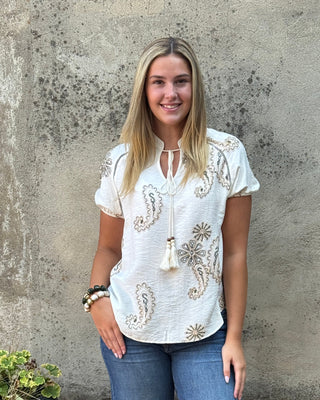 A woman stands outdoors against a textured concrete wall wearing a white short-sleeve bohemian-style top with gold paisley embroidery, blue wide-leg jeans, and brown slide sandals.