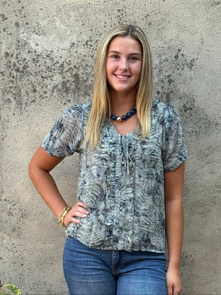 A woman stands outdoors against a textured concrete wall wearing a short-sleeve blue-gray patterned blouse and blue jeans, with a black planter on the left.