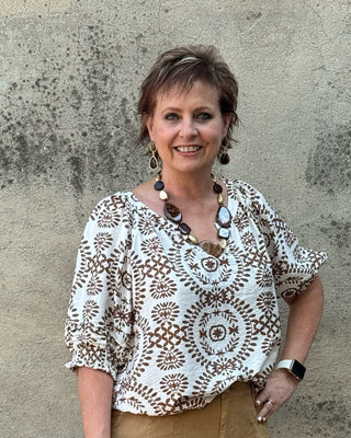 A woman stands outdoors against a textured concrete wall, wearing a bohemian-style short-sleeve blouse with a brown and beige circular mandala pattern on a white background, paired with wide-leg tan trousers and silver sandals.