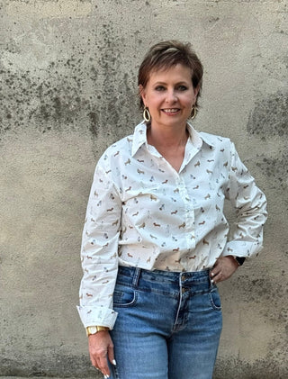 A woman with short brown hair stands against a textured concrete wall, wearing a white long-sleeve button-up shirt with a small brown dog print, tucked into blue jeans.