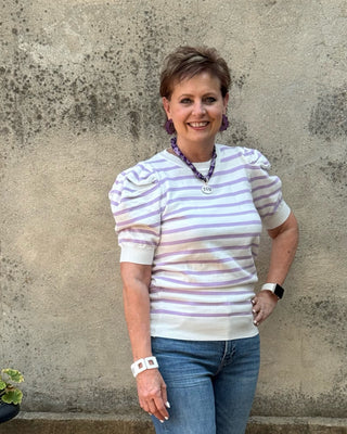 A woman stands outdoors against a textured concrete wall, wearing a white short-sleeve striped top with ruffled shoulders, blue jeans, and white sneakers.