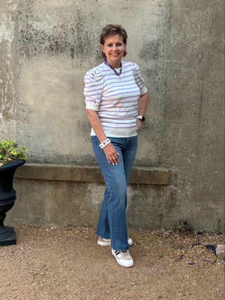 A woman stands outdoors against a textured concrete wall, wearing a white and light blue horizontally striped short-sleeve top with ruffled shoulders, blue jeans, and white sneakers.