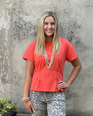 A woman stands outdoors against a textured concrete wall wearing a coral orange short-sleeve peplum top and gray leopard-print cropped pants, accessorized with a chunky gold necklace and bracelets.