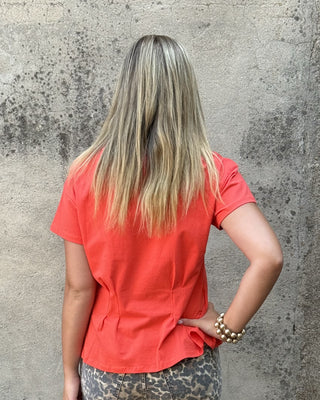 A woman is shown from behind wearing a coral orange short-sleeve top and leopard-print pants, standing in front of a textured concrete wall next to a black planter.