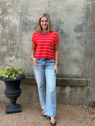 A woman stands outdoors against a textured concrete wall wearing a red short-sleeve top with thin white horizontal stripes, light-wash wide-leg jeans, and brown sandals.