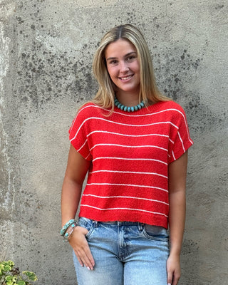 A woman stands outdoors against a textured concrete wall wearing a red short-sleeve knit top with thin white horizontal stripes, light-wash blue jeans, and brown sandals.