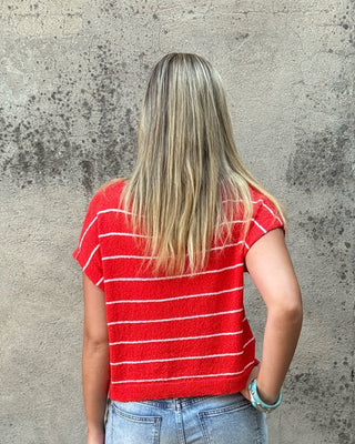 A woman viewed from behind wearing a red short-sleeve t-shirt with thin white horizontal stripes, light-wash blue jeans, and dark shoes, standing in front of a textured concrete wall.