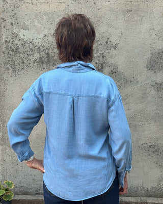 A person viewed from behind wearing a light blue long-sleeve button-up shirt and dark blue jeans, standing against a textured concrete wall.