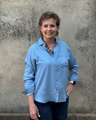 A woman with short brown hair stands against a textured concrete wall, wearing a light blue button-up shirt and dark blue jeans.