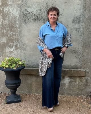 A woman stands outdoors against a textured concrete wall, wearing a light blue button-up shirt with leopard-print sleeves, dark wide-leg jeans, and brown shoes. A black planter with green leaves sits to her left.