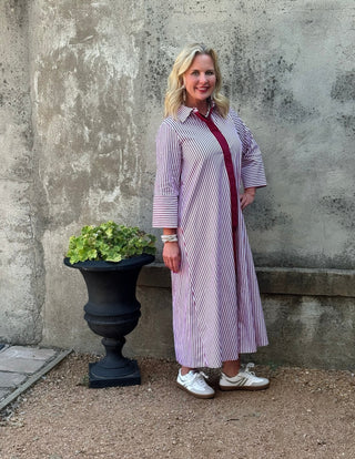 A woman stands outdoors against a textured concrete wall next to a black planter, wearing a lavender and white striped long-sleeve shirt dress with a red tie detail and white sneakers.