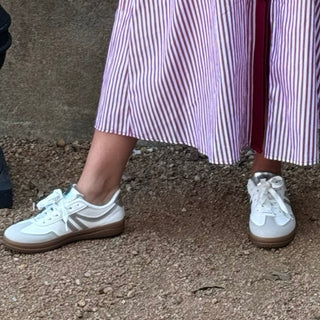 A woman stands outdoors against a textured concrete wall wearing a long-sleeved, button-front midi shirt dress with vertical pink and white stripes and a solid burgundy front placket. She is wearing white sneakers and standing next to a black planter with green leaves.