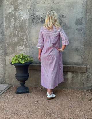 A woman stands with her back to the camera wearing a lavender and white vertically striped midi dress with three-quarter sleeves, posing in front of a textured concrete wall next to a black planter.