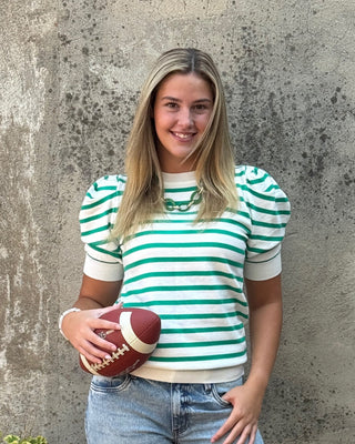A woman stands outdoors against a textured concrete wall, wearing a green and white striped short-sleeve top, light-wash wide-leg jeans, and white sneakers. She is holding a brown football, and a black planter with greenery sits to her left.