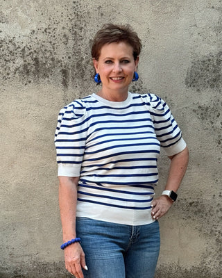 A woman stands against a textured concrete wall wearing a navy and white horizontally striped short-sleeve knit top, blue jeans, and white sneakers.