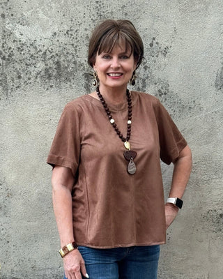A woman wearing a brown short-sleeve top and blue jeans stands in front of a textured concrete wall next to a black planter.