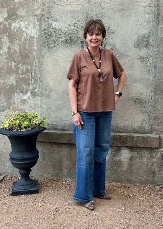 A woman stands outdoors against a textured concrete wall wearing a brown short-sleeve top and wide-leg blue jeans, with a long beaded necklace and leopard-print shoes.