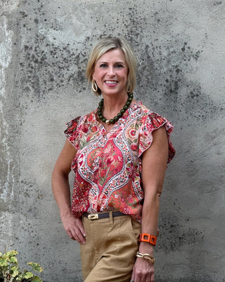 A smiling blonde woman stands against a textured gray concrete wall, wearing a short-sleeve ruffled blouse with a red, orange, and beige paisley pattern, tan pants, and gold jewelry.