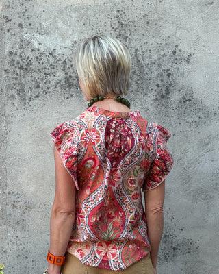 A woman viewed from behind wearing a short-sleeve bohemian paisley blouse with red, pink, and green patterns, paired with tan pants, standing against a textured concrete wall.