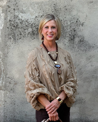 A smiling blonde woman wearing a beige ruffled blouse, layered necklaces, and a gold watch, standing against a textured concrete wall.