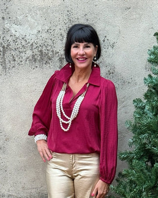 Woman in a red blouse and gold pants standing next to a potted tree against a concrete wall.