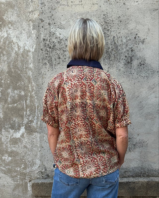 A person viewed from behind wearing a short-sleeve button-up shirt with a rust-orange and beige geometric floral pattern and a dark navy collar, paired with blue jeans, standing against a textured concrete wall.