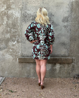 A woman with blonde hair is seen from behind, wearing a short puff-sleeve dress with a brown, white, and turquoise floral pattern, standing in front of a textured concrete wall.