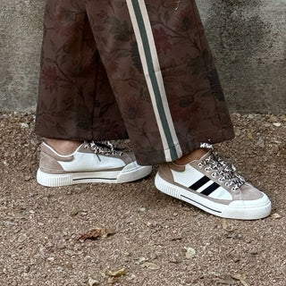 A woman posing outdoors in front of a textured concrete wall, wearing a brown faux-leather tracksuit with cream stripes and white sneakers.