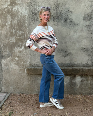 A woman posing outdoors in front of a textured concrete wall, wearing a cream, orange, and black striped long-sleeve top, blue wide-leg jeans, and white sneakers.