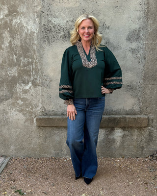 blonde haired woman in a green top with cheetah pint details and jeans standing in front of concrete wall