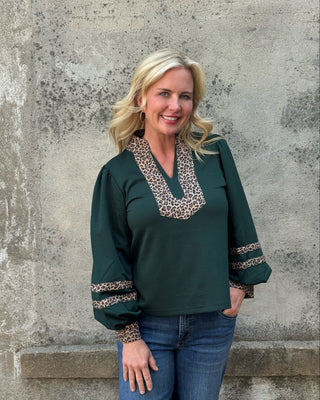A woman with blonde hair wearing a dark green long-sleeve blouse with leopard print trim on the V-neck and cuffs, paired with blue jeans, standing against a textured concrete wall.