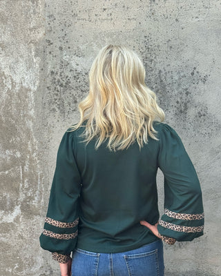 A woman viewed from behind wearing a dark green satin blouse with leopard-print trim on the cuffs, paired with blue jeans, standing against a textured concrete wall.
