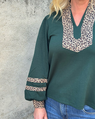 A woman wearing a dark green long-sleeve top with leopard print trim on the V-neck and cuffs, paired with blue jeans, standing against a textured gray concrete wall.