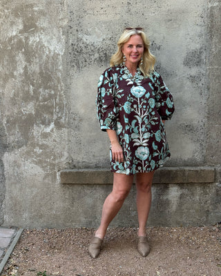 A woman stands against a textured concrete wall wearing a short-sleeved button-up dress with a dark brown base and a light blue floral pattern, paired with taupe pointed-toe flats.