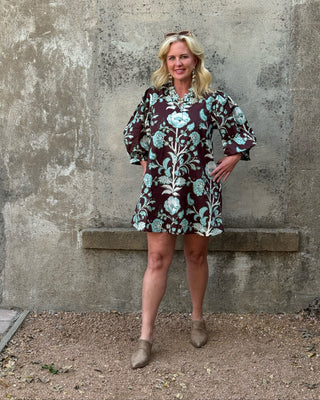 A woman stands outdoors against a textured concrete wall, wearing a short floral print dress with long puff sleeves and taupe ankle boots.
