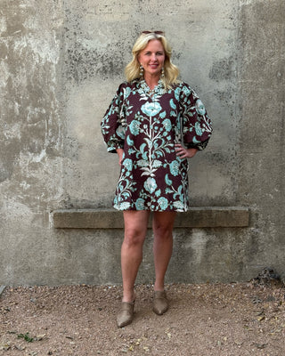 A woman stands outdoors against a textured concrete wall wearing a short-sleeve button-front dress with a dark burgundy base and a light blue and white floral pattern. She is wearing brown ankle boots.