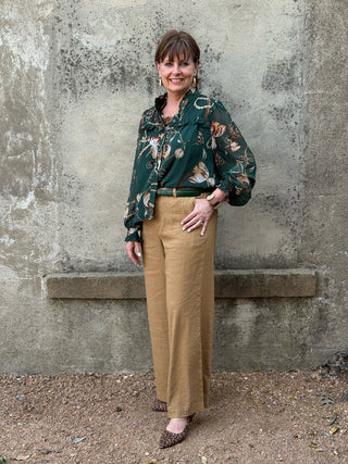 woman standing in front of a concrete wall in a green silky blouse and tan linen pants