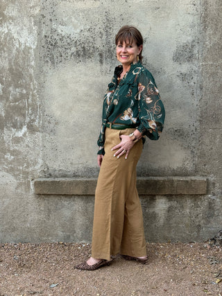 woman standing in front of a concrete wall in a green silky blouse and tan linen pants
