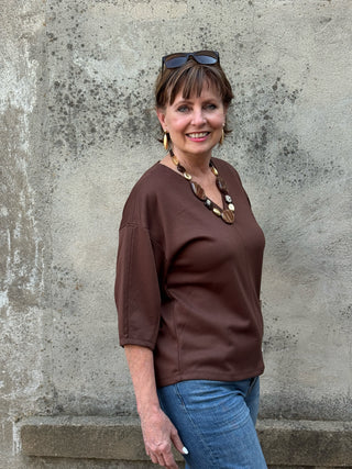 A smiling woman with sunglasses on her head stands against a textured concrete wall, wearing a brown V-neck top and blue jeans.