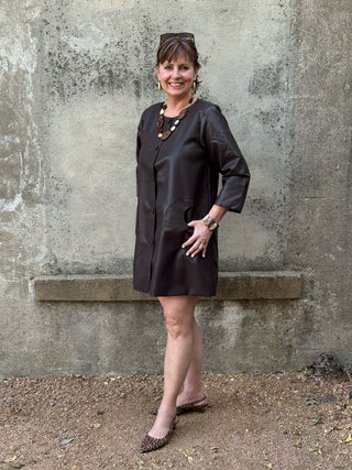 A woman modeling a dark brown short-sleeve button-up dress with a relaxed fit, standing against a textured concrete wall.