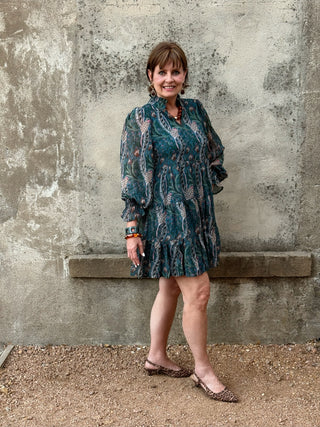 A woman stands outdoors against a textured concrete wall wearing a teal and brown patterned long-sleeve mini dress with ruffled trim, paired with leopard-print flat shoes.