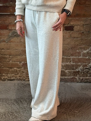 Woman in a white tracksuit standing in front of a fireplace with decorative plants on a brick wall.